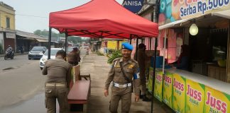Dua petugas Satpol PP Cilegon berjaga di bawah tenda merah di trotoar Pasar Kranggot, berdiri di dekat warung minuman jus.